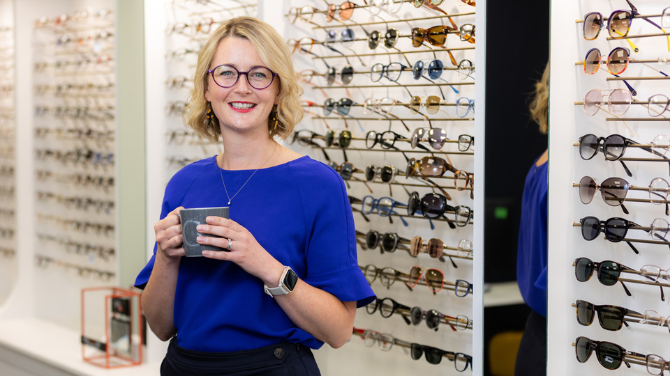 Woman standing in an optical practice with short blonde hair wearing a bright blue shirt and glasses. She is holding a mug in both hands and smiling