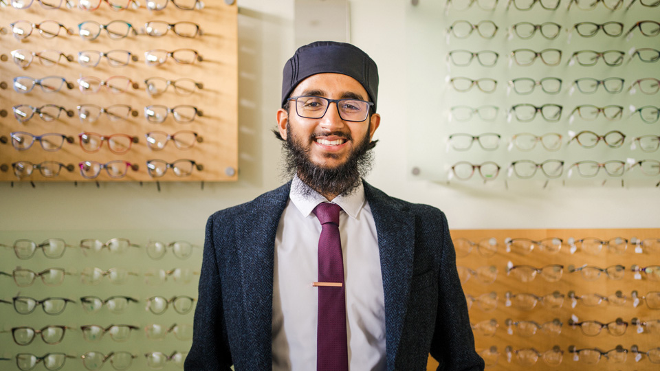 Habeeb is smiling and is stood in front of an orange and green wall which is displaying a large number of spectacle frames 