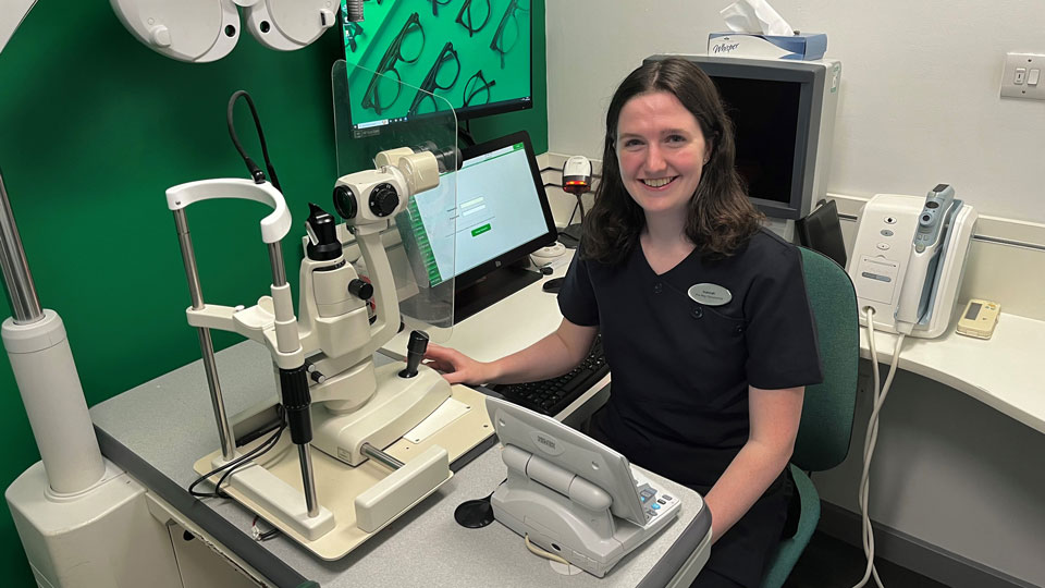 Hannah is smiling whilst sitting at a table next to a green wall in a Specsavers testing room. A large piece of optical equipment is on the table in front of her