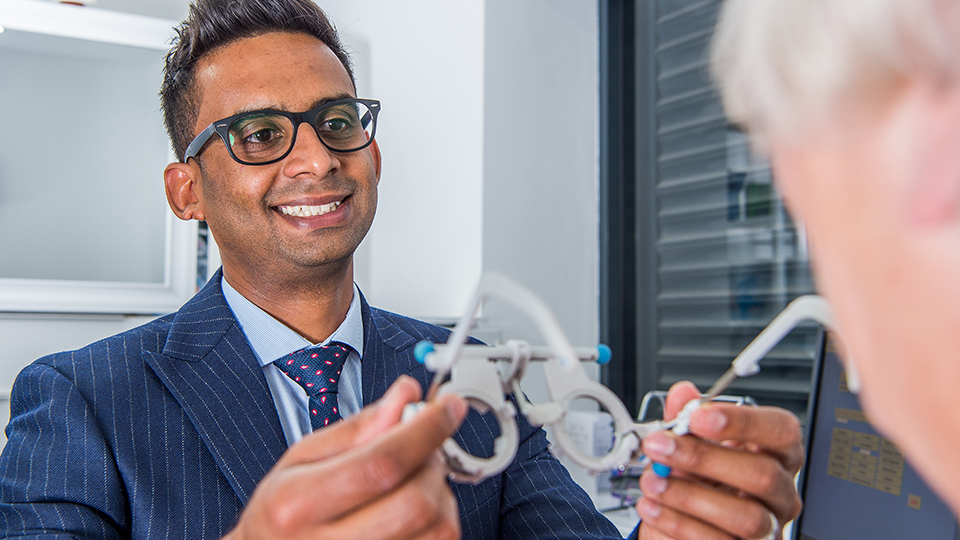 Harjinder Sunda is in practice, holding a trial frame towards the camera and towards a patient whose shoulder can be seen 