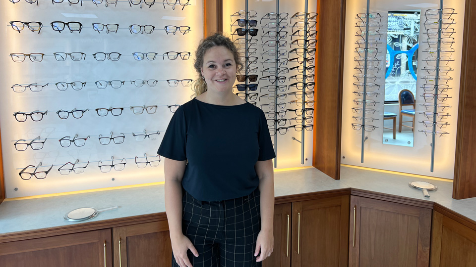 Harriet stands in an optometry practice before wooden and mirrored wall displays of spectacles and sunglasses