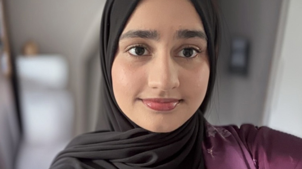 Taking a selfie, Hifsah is smiling softly. She wears a warm purple top and black headcovering