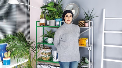 A woman in a grey turtleneck and jeans stands in front of a backdrop of pot plants on shelves 
