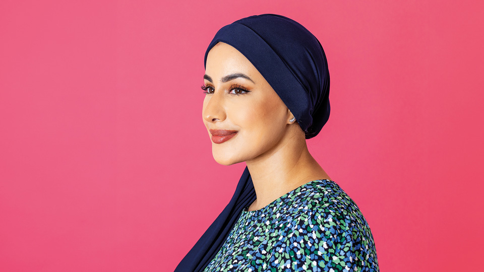 Profile of a female wearing a dark blue head scarf and a blue top on a bright pink background