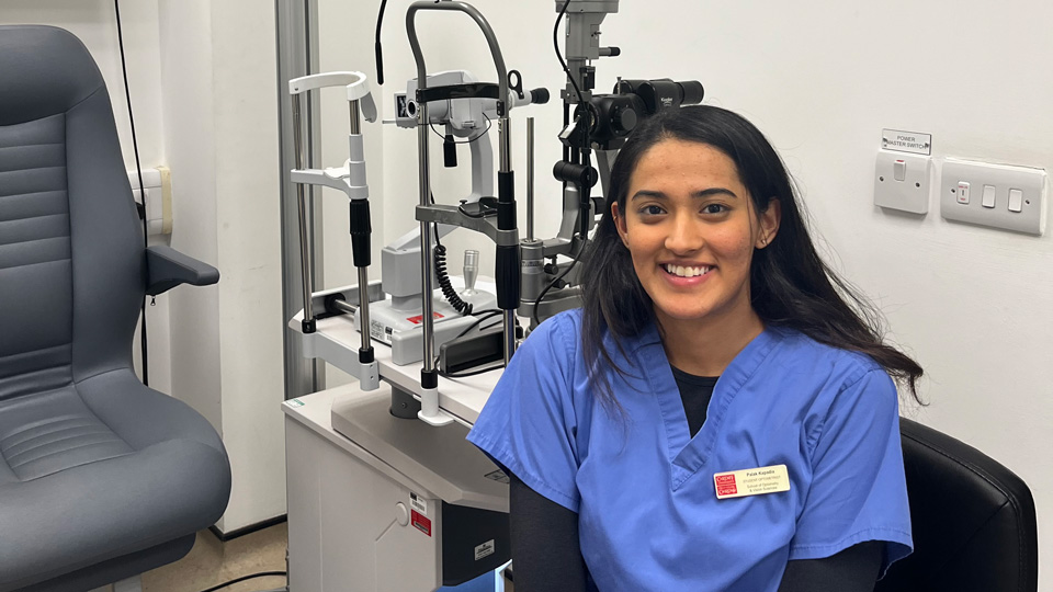 Palak wears blue medical scrubs and is seated in a clinic setting in front of a range of instruments including a slit lamp. Against one wall is the test chart 