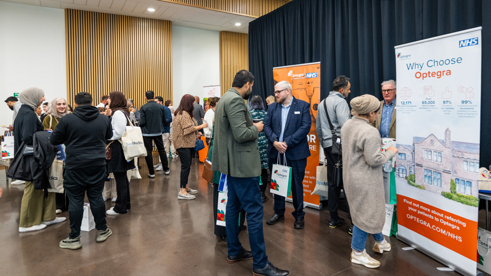 Optometrists gather in a university hall holding bags with sponsor and exhibitor branding, around the room are exhibitors with tall banners