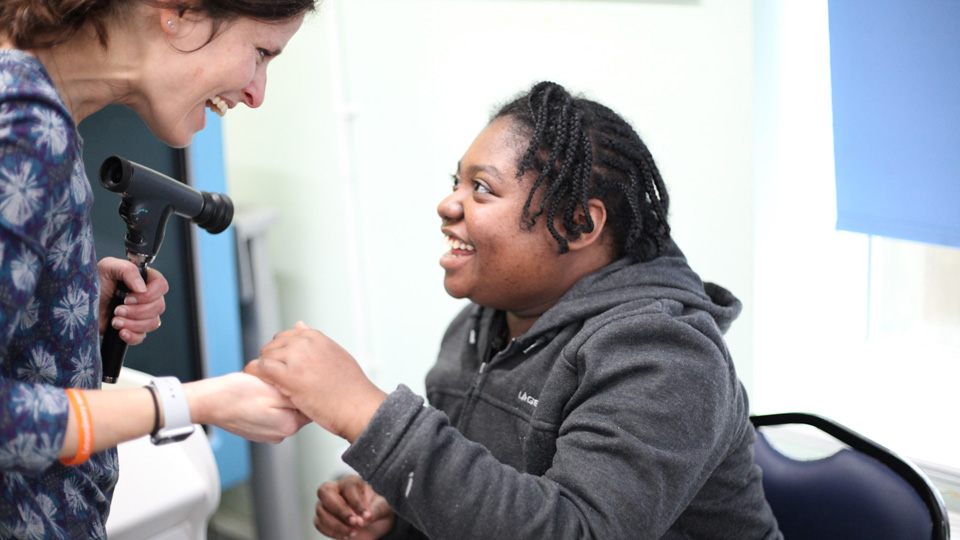 Lisa is conducting a sight test, holding in one hand an instrument, in the other she holds the hand of Abigal. They are looking into each other’s eyes and smiling broadly 