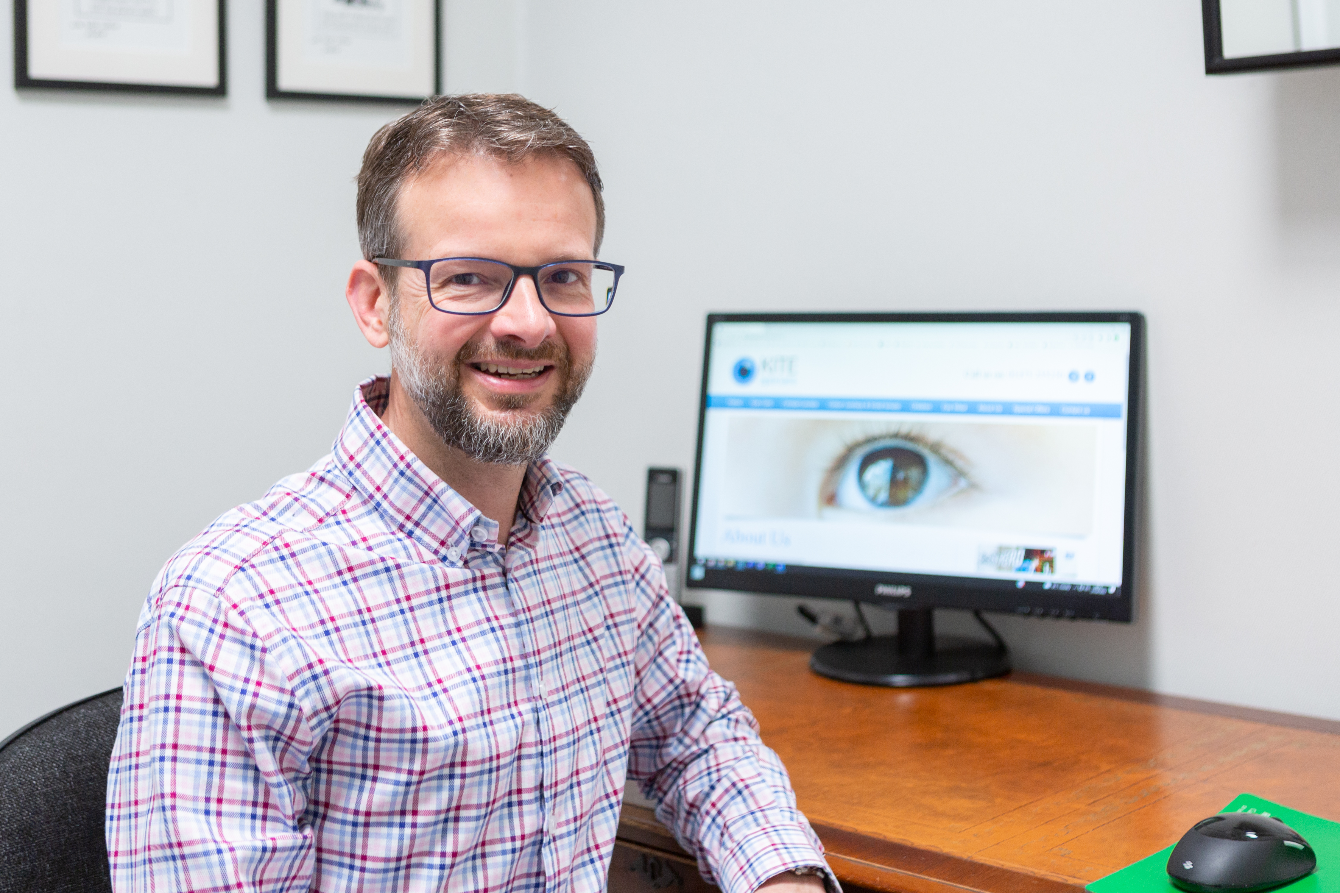 Adrian is seated at a desk in practice. The screen behind is out of focus but displays the website of Kite Opticians