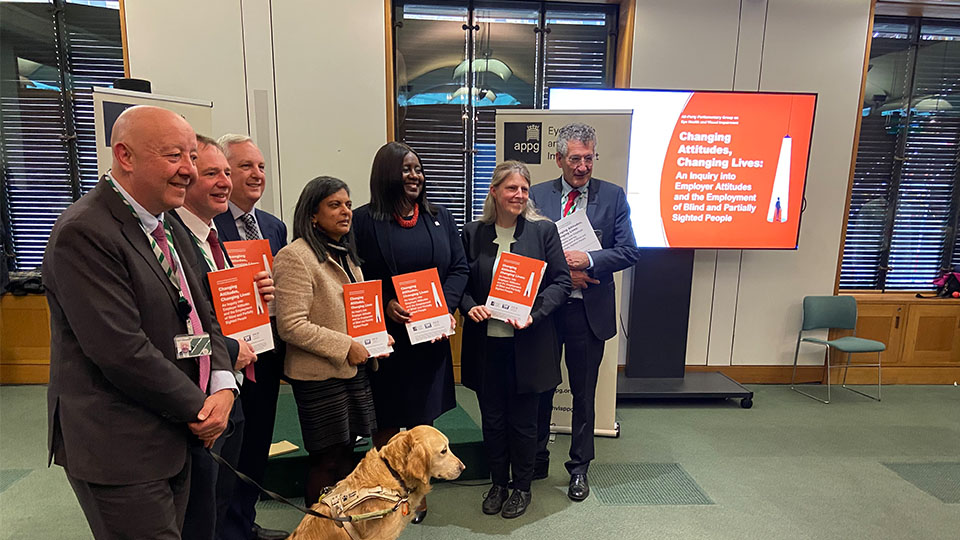 Seven people standing side by side, all dressed in smart clothes A few people are holding a orange A4 sized leaflet. An assistance guide dog is sitting in front of the group 