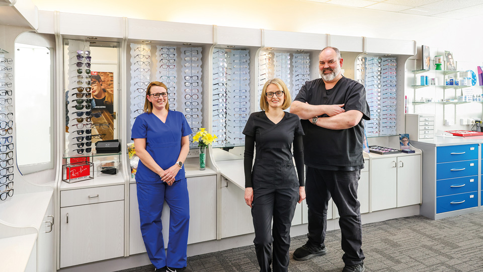 Three smiling staff wearing scrubs stand on the floor of an optometry practice. Behind them are display cabinets with spectacle frames and a vase with daffodils. The woman on the left is wearing blue scrubs, spectacles and a watch. The woman in the centre is wearing spectacles and black scrubs over a long-sleeved black top. The man on the right is wearing black scrubs, a watch and has his arms crossed in front of him. 