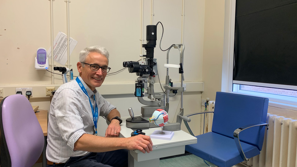 Chris Steele is sat at a desk in his consulting room, smiling at the camera, with a piece of optical testing equipment on the desk in front of him 