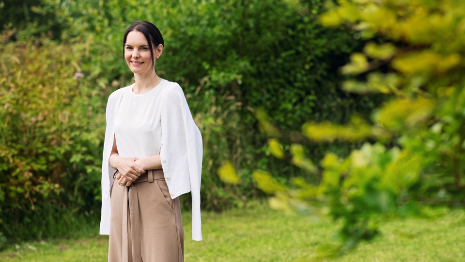 Christina is stood next to a leafy green tree in a garden, wearing beige trousers and a white cardigan 