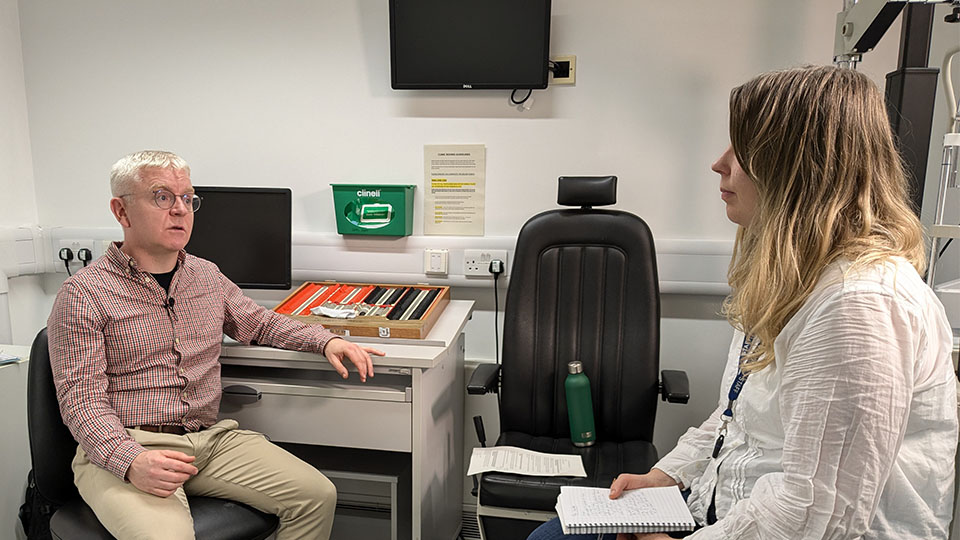 On the left a male optometrist is sitting in a practice testing room and on the right is a female journalist holding a notepad.  