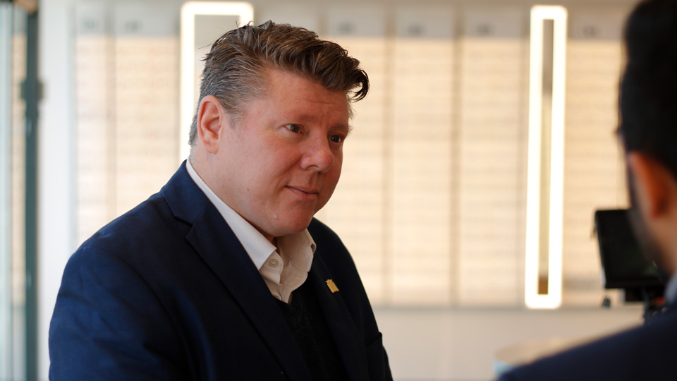 Dean Russell is wearing a dark blue suit and speaking to someone to the right of the camera lens  