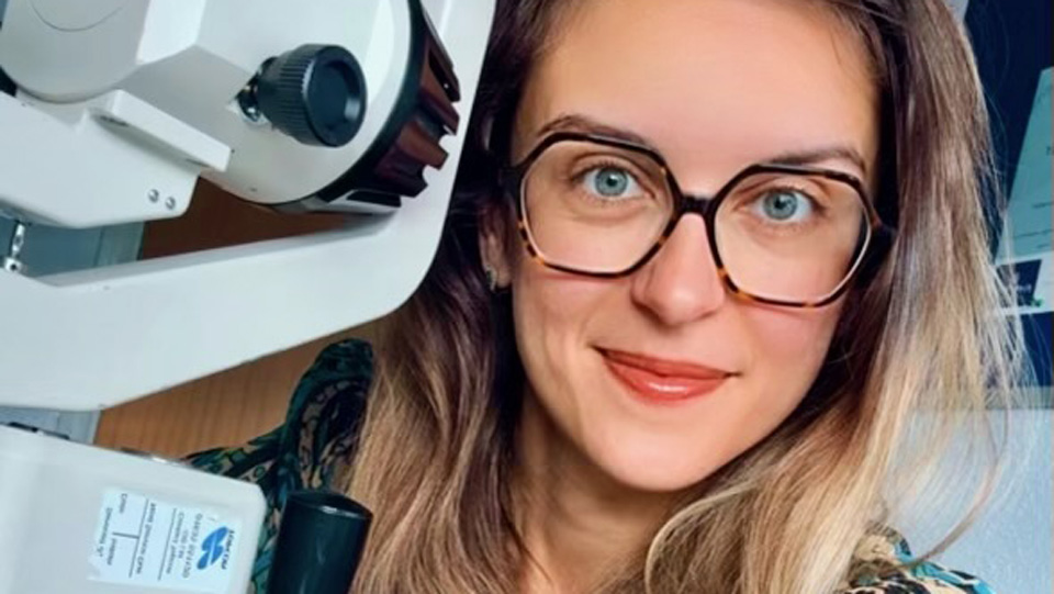 Desislava smiles while posing next to a piece of optical equipmernt in a testing room. She is wearing large framed glasses