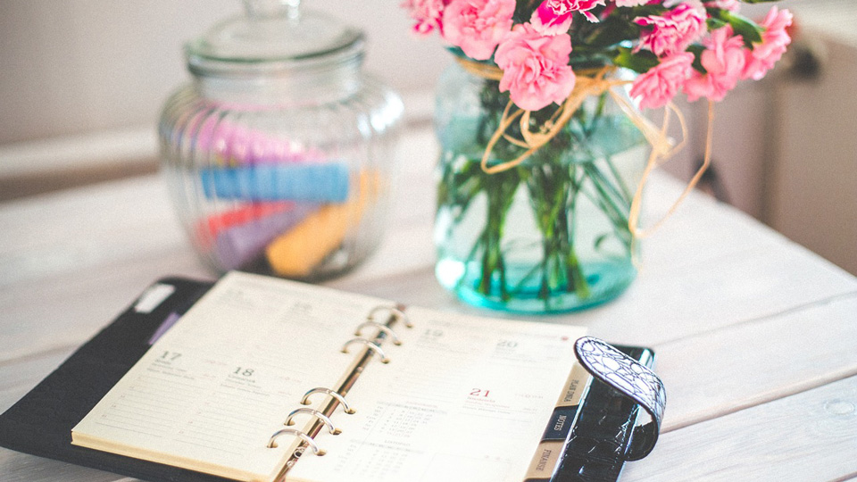 A diary sits on a white table net to a vase of pink carnations and a glass jar containing different coloured pieces of chalk  