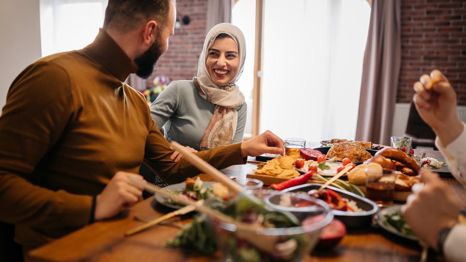 A woman wearing a light hijab smiles at a man in a brown turtleneck jersey. They are eating lunch with a selection of different dishes on the table 