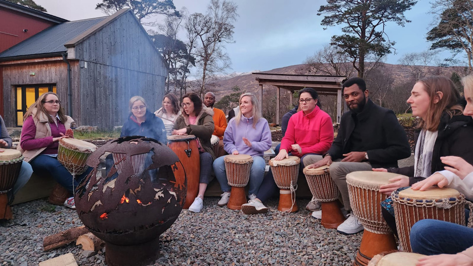 Attendees are sat around a fire pit playing drums, with a mountain in the background
