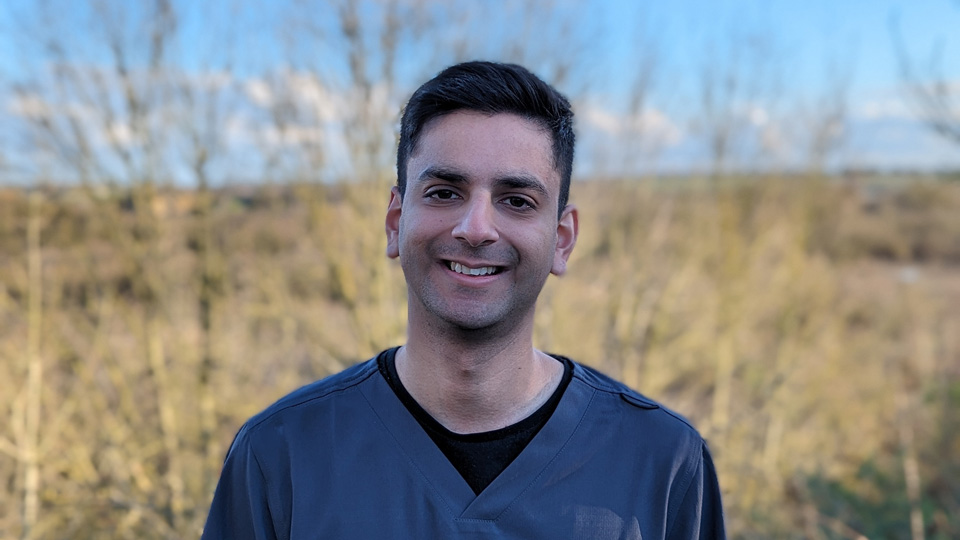 Karan is smiling in front of a grassy field and is wearing dark blue medical scrubs  