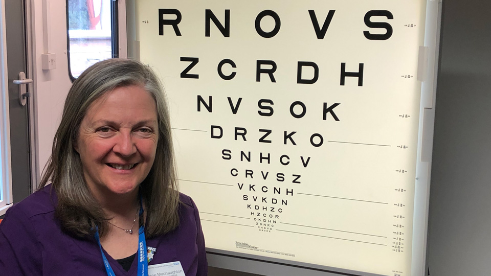 A woman in a purple t-shirt with a blue lanyard around her neck. She is standing next to a large Amsler chart. 