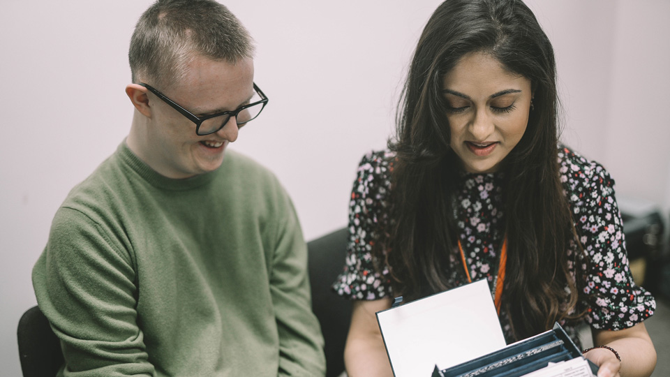 Janki is looking down at a screen whilst a patient in glasses and a green jumper sits next to her smiling  