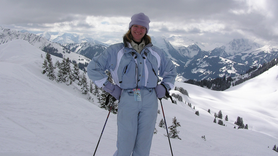A woman in a light blue ski suit is smiling in the snow up a mountain 