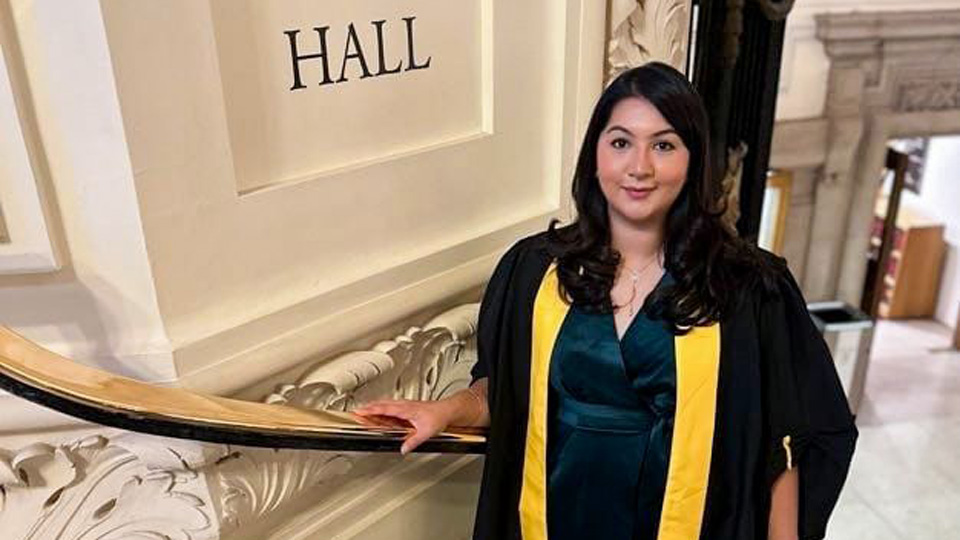 Kamal is dressed in a black graduation gown with a yellow stripe, and is standing on stone steps inside a grand building  