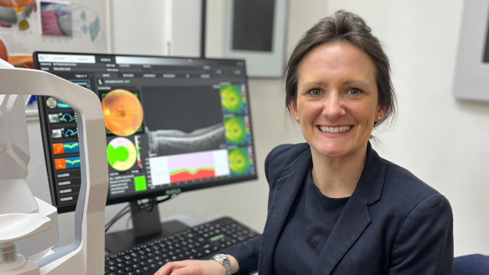 Katherine wears a formal dress suit and is sat in a consulting room in front of a computer screen displaying retinal images. The wall has a large anatomical poster of the eye. 