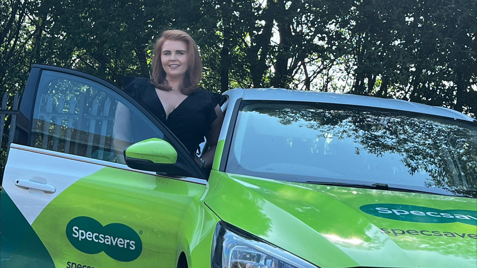 A young woman with red hair is standing behind the open door of a lime green, Specsavers branded car