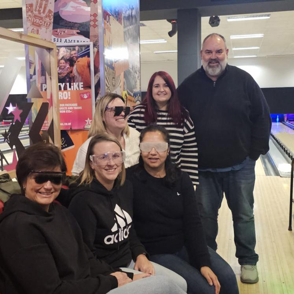 A team of people gather in a bowling alley, several wear goggles of varying shades of visibility 
