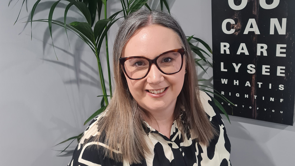 Linda wears bold black spectacles and a black and white top, standing in front of an artwork that mimics the snellen chart