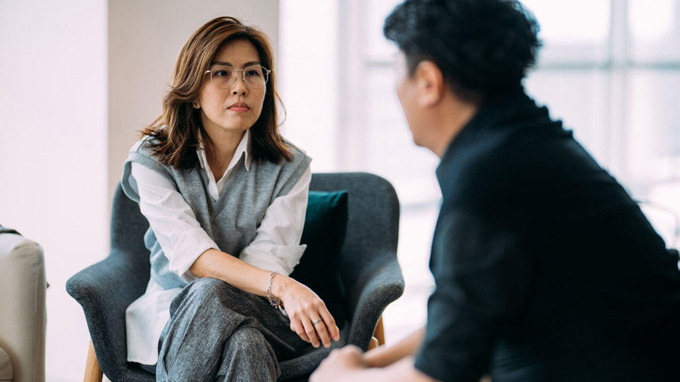 A lady sitting on a chair, legs crossed, opposite a man sitting on a chair, hands crossed, looking at each other