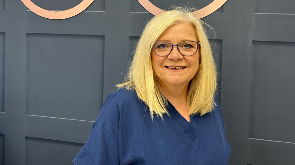 Lynn poses in navy scrubs against a sign reading Eyeglass Opticians
