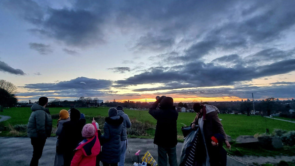 A crowd of people wearing coats and woolly hats gather close to a field. A girl in a red jacket and pink hat in the centre of the crowd is pointing at the sky. There is an orange stripe of light low across the horizon as the sun sets with darker clouds moving across a light grey sky.   
