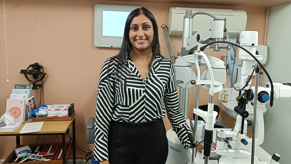 Nikita stands in a testing room, which has wall painted a calming peach, and an array of optical equipment. She wears a black and white shirt which has stripes going in lots of directions. 