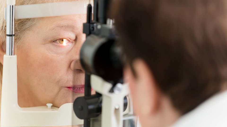 Over the shoulder of an optometrist who looks through the lens of a slit lamp, an older woman is having a light shone into her eye for applanation tonometry