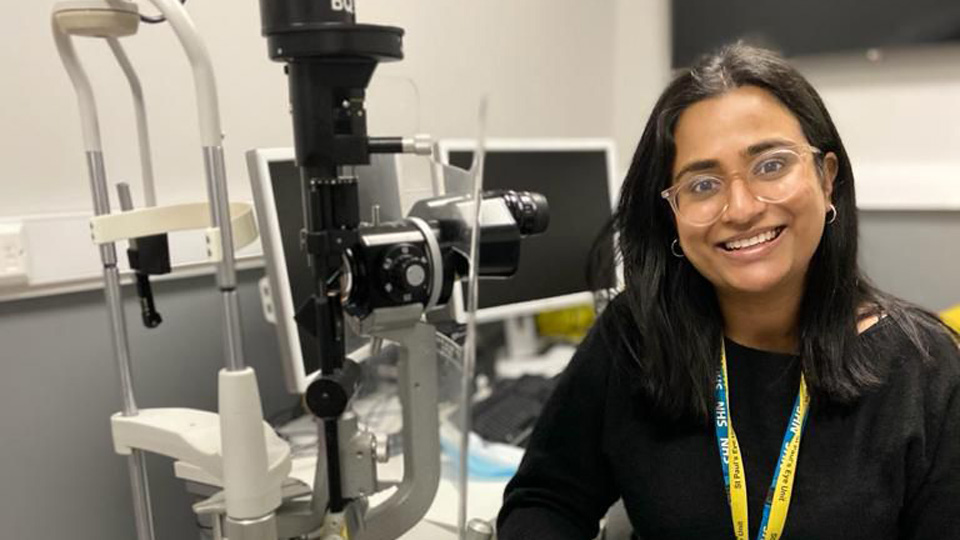 Rupa is wearing a black top and green skirt, and is smiling whilst sat at a desk that holds optical equipment 