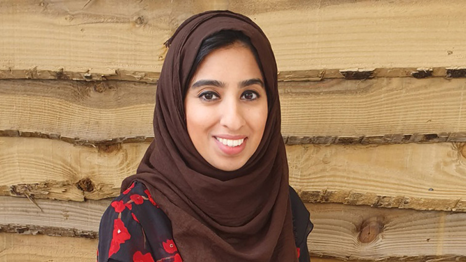 A woman wearing a brown hijab and dark top with a red pattern smiles at the camera 