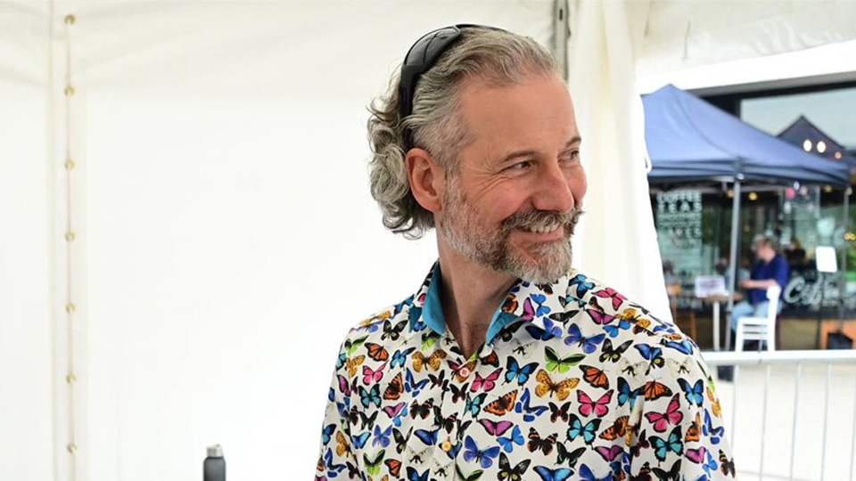 Simon is looking to the left of the camera and is wearing a white shirt decorated with multicoloured butterflies 