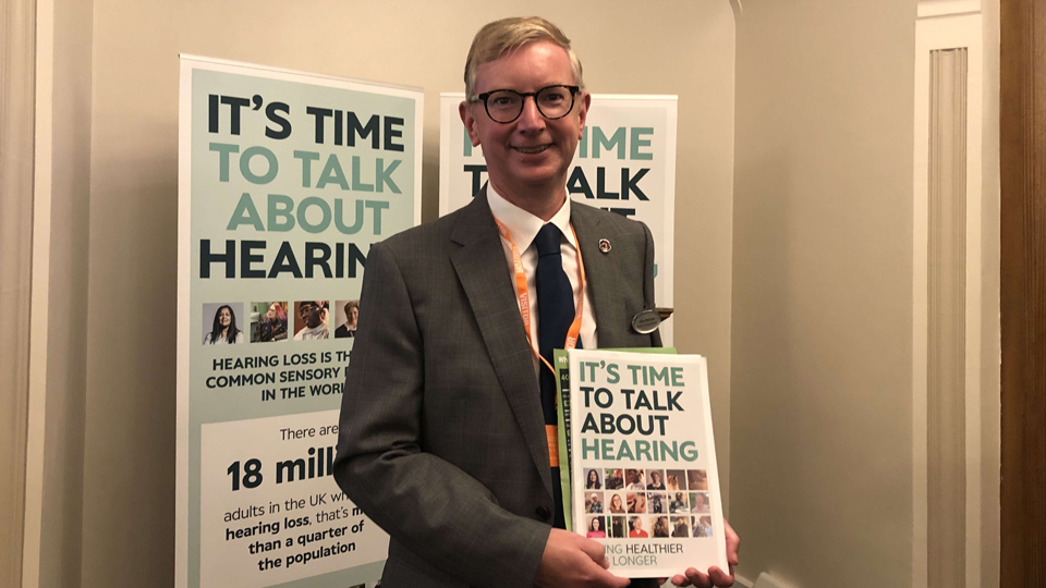 A man with black framed glasses wearing a suit is holding a A4 sized with a title that reads, It's time to talk about hearing