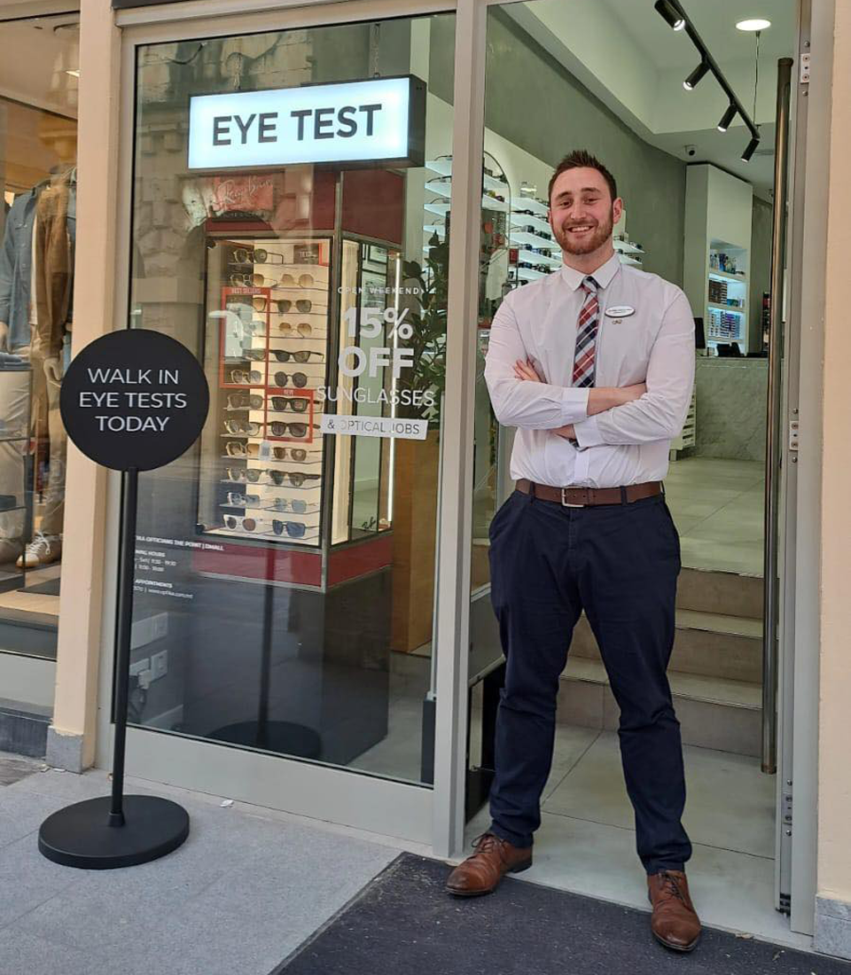 A young man, Luke, standing with his arms crossed and smiling in front of an optometry practice. He is smartly dressed with dark blue trousers, a white shirt and tie. 