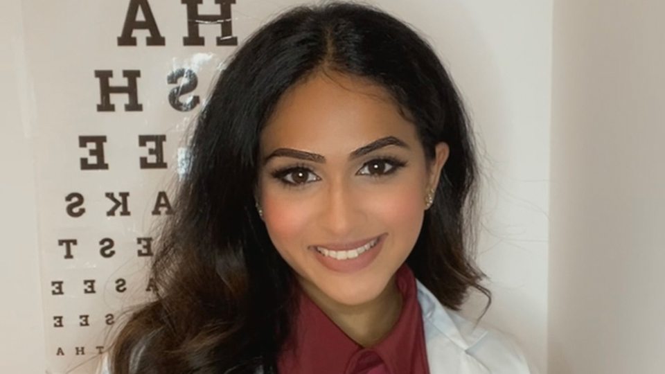 A South Asian girl with long black hair and a white lab coat is smiling in front of an Amsler chart 