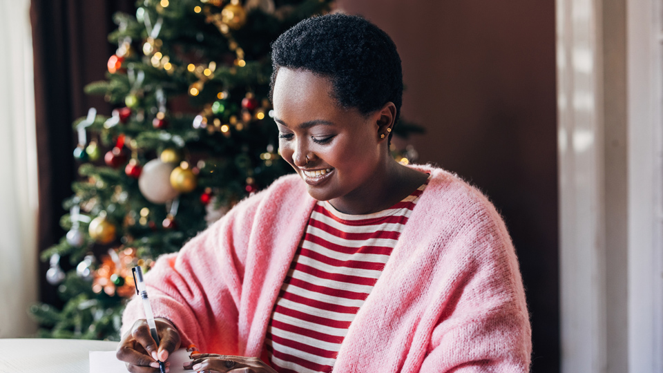 A woman in a pink jersey and striped top writes a list with a christmas tree in the background