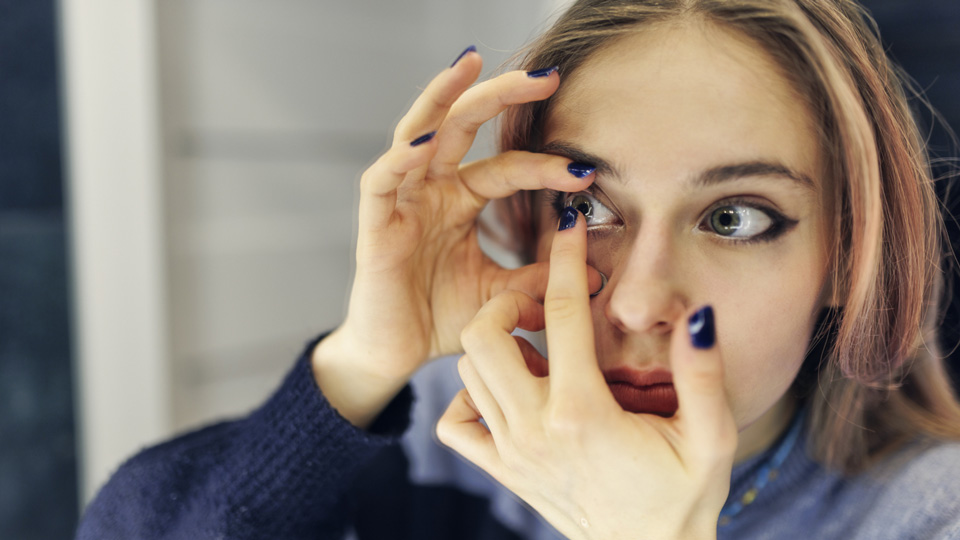 A young woman with light brown hair is applying a contact lens in front of a mirror 