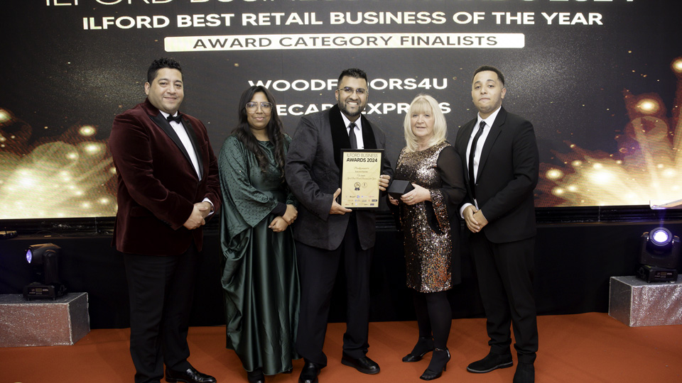 Five people stand in front of a display for Ilford Business Awards. Everyone is dressed in smart suits and dresses in navy, black, green, and deep red. 