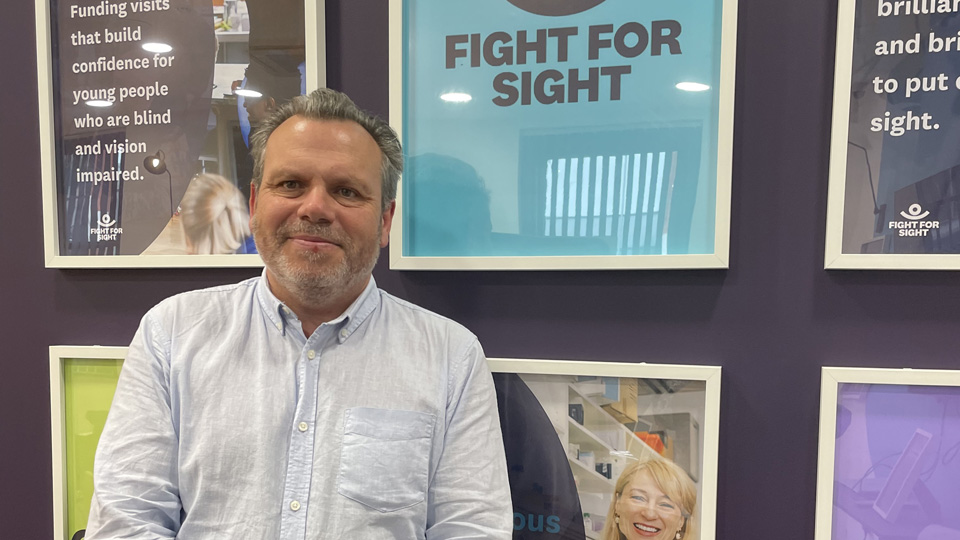 Keith Valentine is stood in front of a navy blue wall that displays posters and the new turquoise Fight for Sight branding  