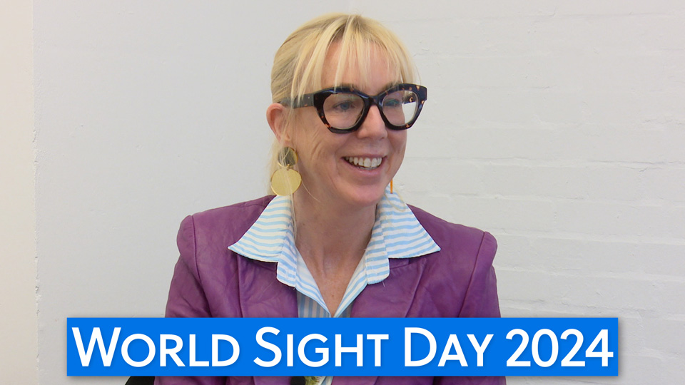 A still from a video interview with Caroline Casey who wears oversized chunky black frames, a purple leather-style jacket, and blue striped shirt with palm trees