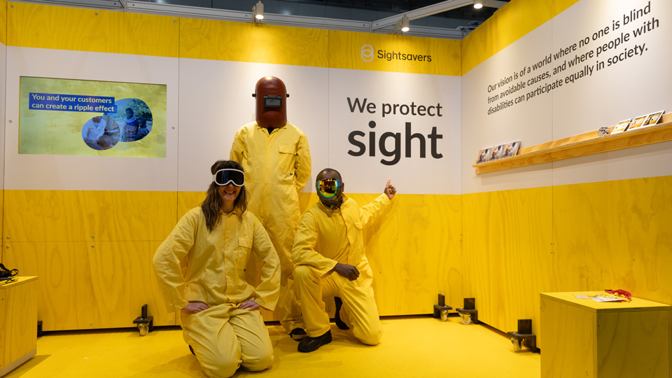 Three colleagues wear bright yellow protective clothing and kneel down inside the Sightsavers exhibition stand 
