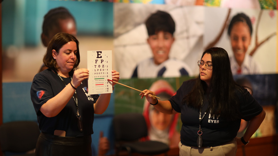 Two eye care professionals wearing Love Your Eyes branded t-shirts hold up a Snellen chart