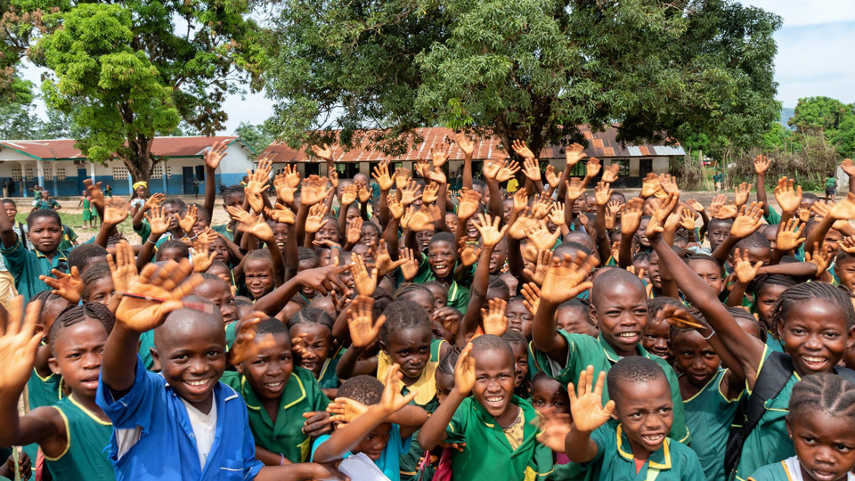 School children are gathered, smiling and waving at the camera